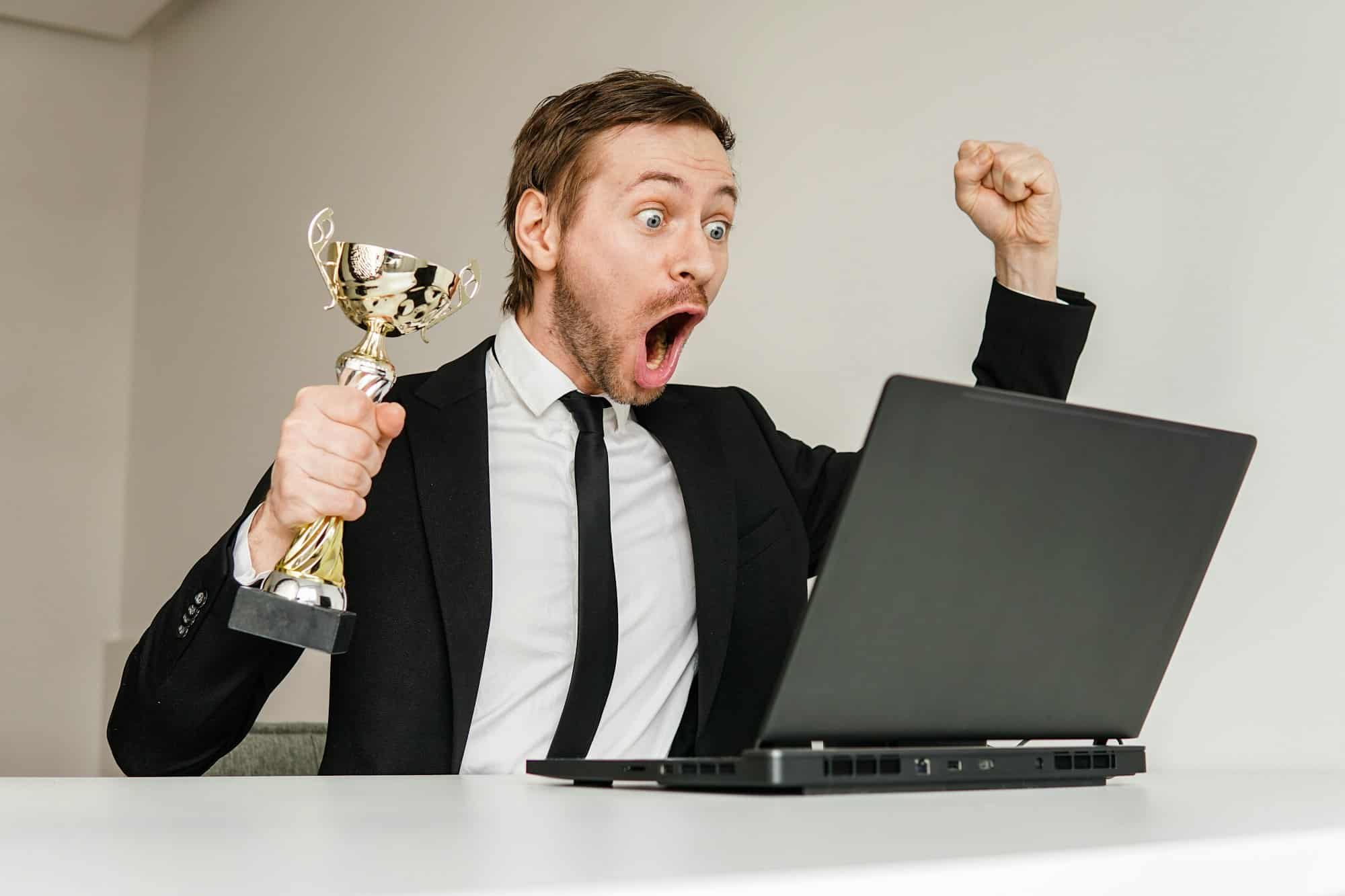 Successful businessman celebrating victory with cup trophy while watching online ceremony on laptop.